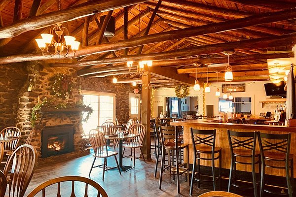 A cozy restaurant interior with a fireplace and a bar counter with stools. There are several tables and chairs in the background. This restaurant might be Stone House Tavern located in Accord, New York.