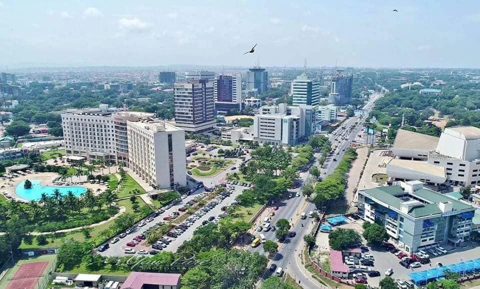 An aerial view of a busy Accra Ghana city street with heavy traffic. Cars, buses, and taxis can be seen moving along the street. Tall buildings line either side of the street.