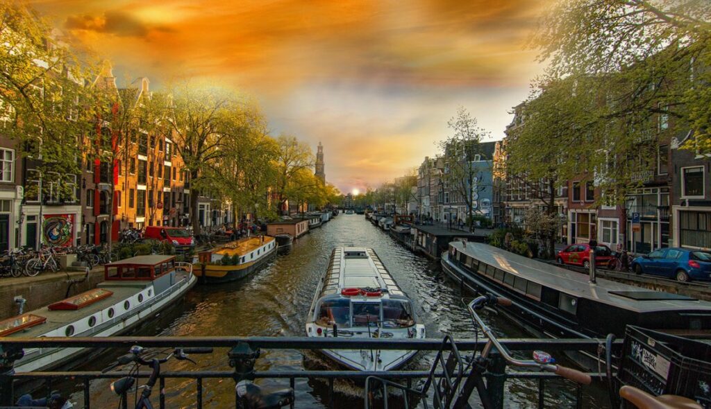 A photo of a narrow canal with a small tour boat traveling down it. The canal is lined with buildings on either side, some of which are shops with colorful awnings. There are trees with green leaves lining the canal. This is likely a scene in Amsterdam, Netherlands.