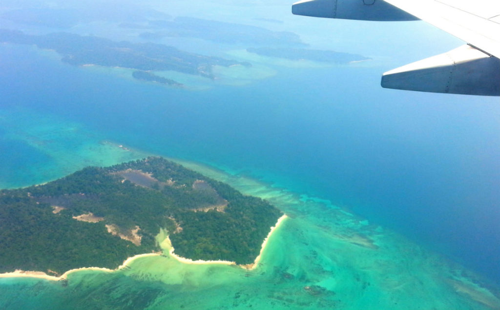 An aerial view of a Andaman And Nicobar island paradise. The island is lush and green, with a white sand beach bordering the turquoise water. The beach is lined with palm trees. In the center of the island, there is a small cluster of buildings with thatched roofs.