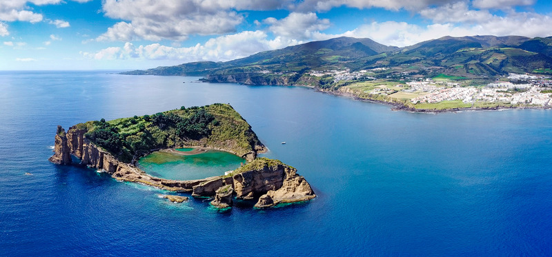 This image is of Azores Portugal with small, rocky island with lush green vegetation in the middle of a vast ocean. The island has steep cliffs and a beach. In the background, there is a range of steep, green mountains.