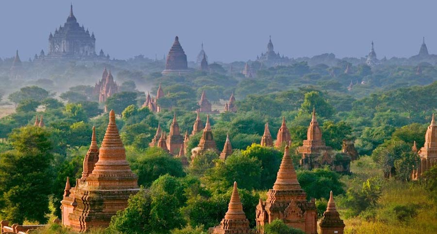 A photo of a cluster of ancient temples made of red sandstone, nestled amongst trees in a lush green forest. In the background, there is a large, modern building. The temples are located in Bagan, Myanmar.
