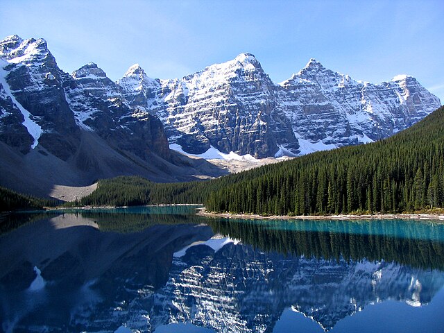 A calm lake reflects the snow-capped peaks of a mountain range. Evergreen trees line the shore of the lake.