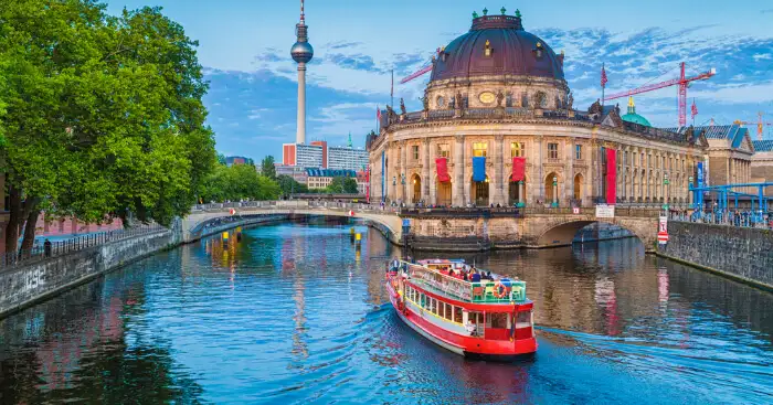 A red and white passenger boat travels down a river in front of a large, classical building with a dome. The building is the Bode-Museum in Berlin, Germany. There are trees with green leaves lining the riverbank.