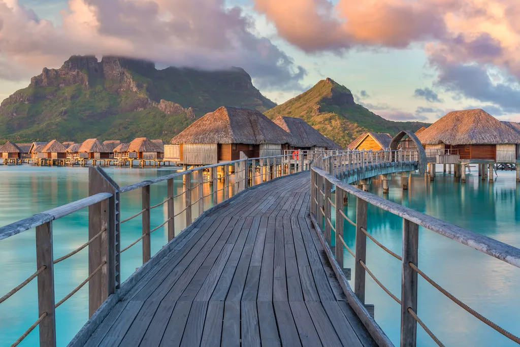 A wooden walkway leads over clear blue water to thatched-roof bungalows on a tropical island. Lush green vegetation surrounds the bungalows. There are mountains in the background.