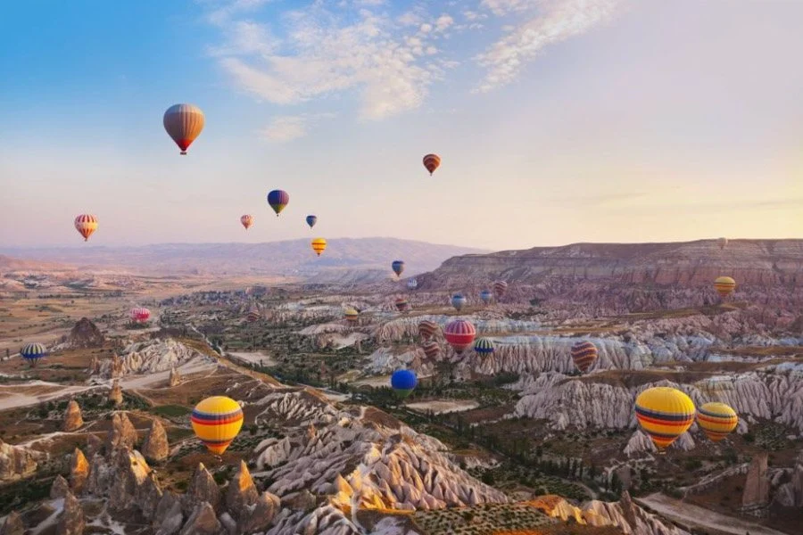 A group of colorful hot air balloons float over a rocky landscape with mesas and buttes. The sky is clear and blue.