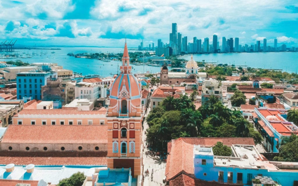 An aerial view of Cartagena, Colombia, with the iconic clock tower in the foreground. The clock tower has a red tiled roof and a clock face on each of its four sides. In the background is the busy city skyline of Cartagena.