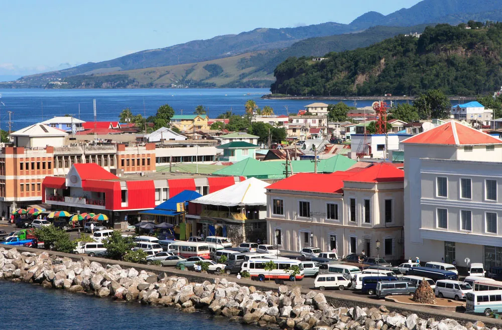 A photo of a Dominica Roseau with colorful buildings lining a street. Cars are parked along the side of the road. The city is located next to a blue ocean.