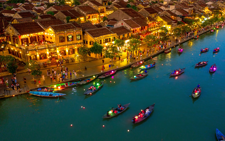 This image is of hoi an vietnam the best place to travel it is A night scene of boats on a river, with city lights reflected on the water.