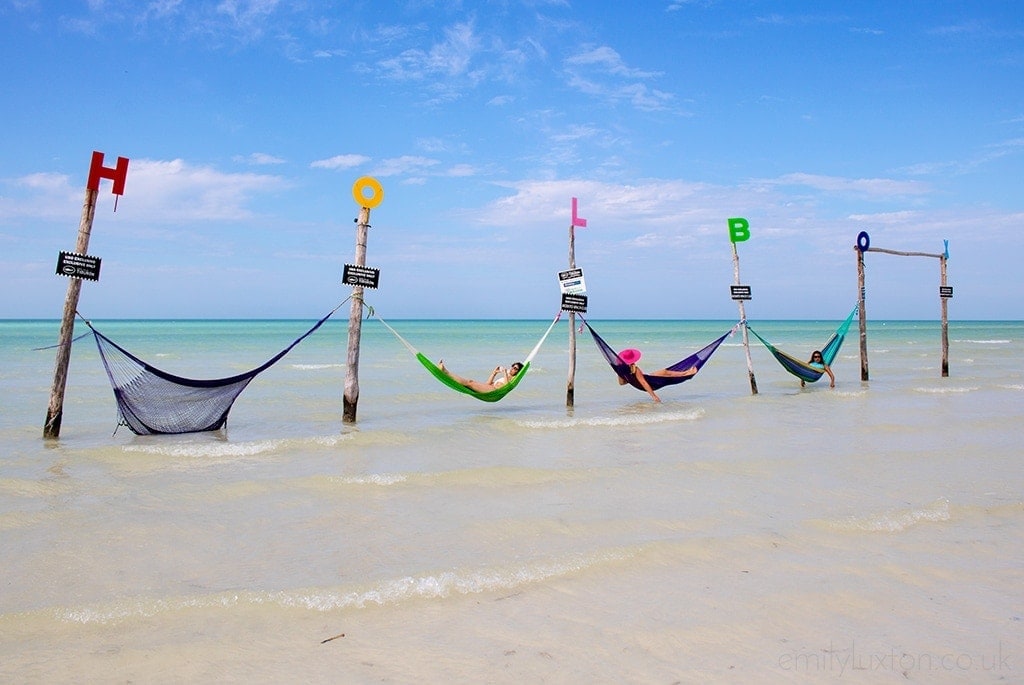 A person relaxing in a hammock strung between two palm trees on a beach at Isla Holbox Mexico. The person is wearing a swimsuit and sunglasses and appears to be enjoying the view. The beach has white sand and clear blue water. There are other palm trees scattered around the beach.