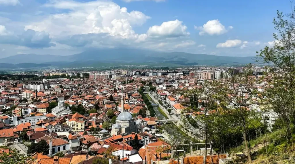 An aerial view of a Kosovo sprawled out from a hilltop in the foreground. The city is filled with buildings in various shapes and sizes. The surrounding landscape includes rolling hills, a river that winds through the city, and a lake in the distance. The sky is a bright blue with some white clouds.