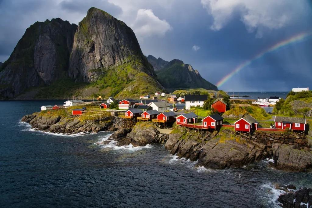 A photo of a cluster of colorful houses with shingled roofs on a cliff overlooking a blue ocean. The houses have balconies and decks facing the water. There is green vegetation growing on the cliffside.