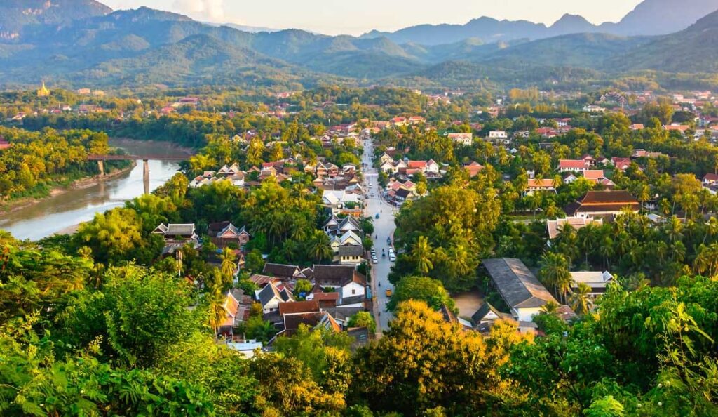 A photo of a small town in Southeast Asia, Luang Prabang Laos, located on a riverbank. The surrounding area is mountainous and lush with vegetation.