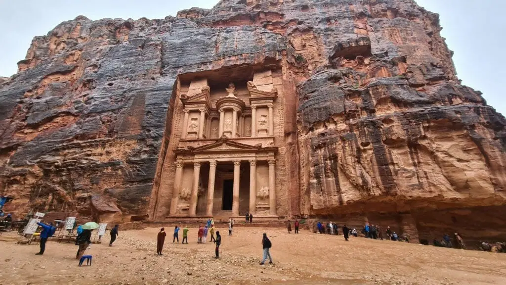 An image of the Al-Khazneh, also known as the Treasury, in Petra, Jordan. It is an elaborate temple carved from the red sandstone cliff face. The Treasury has a wide porch with intricately carved columns and friezes. There is an urn atop the central structure. Tourists are standing in the foreground.