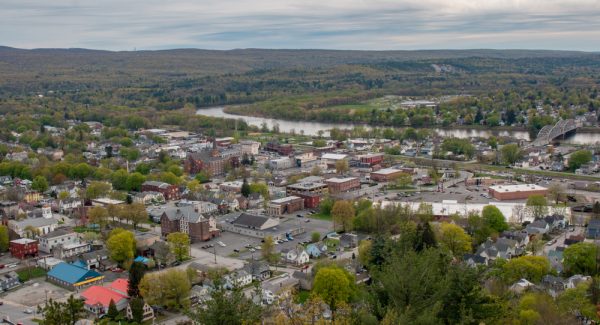 An aerial view of a Port Jervis Newyork nestled amongst a dense forest. The town has a central winding river with a bridge crossing it. The buildings appear colorful and there’s a church with a tall steeple in the center.