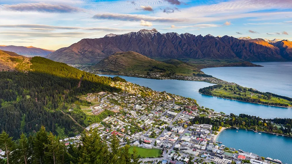 A scenic view of a queenstown NewZealand city nestled between snow-capped mountains and a calm lake. The city has buildings with colorful roofs and a central church with a large dome.