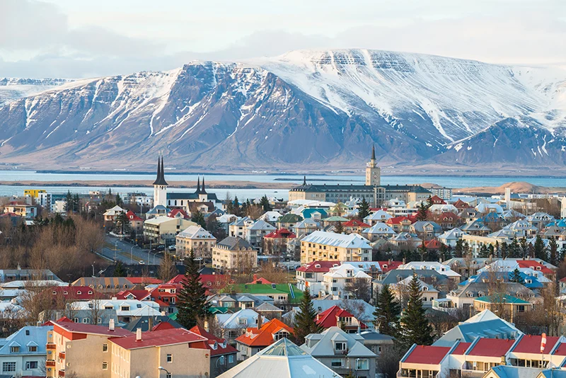 A photo of Reykjavik, Iceland in late winter. The city has colorful buildings with red roofs. Hallgrímskirkja, a large white church with a distinctive tower, dominates the skyline. The mountains in the background are snow-capped.