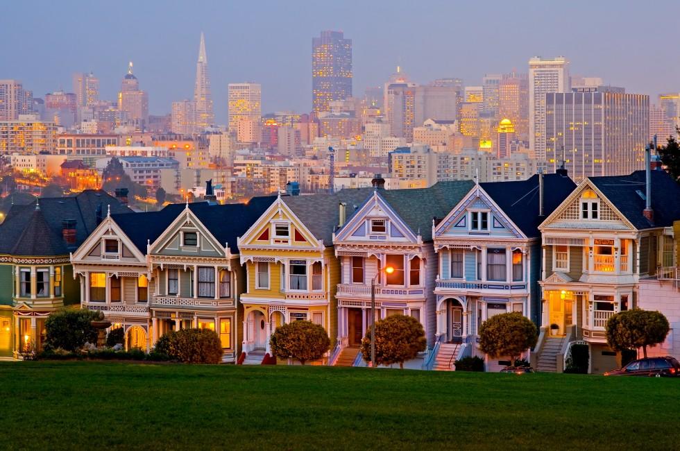 A row of colorful Victorian houses with a city skyline in the background. The houses are painted in bright colors such as red, blue, yellow, and green. Some houses have bay windows and balconies. The city skyline in the background is made up of modern skyscrapers. This is likely a view of Alamo Square in San Francisco.