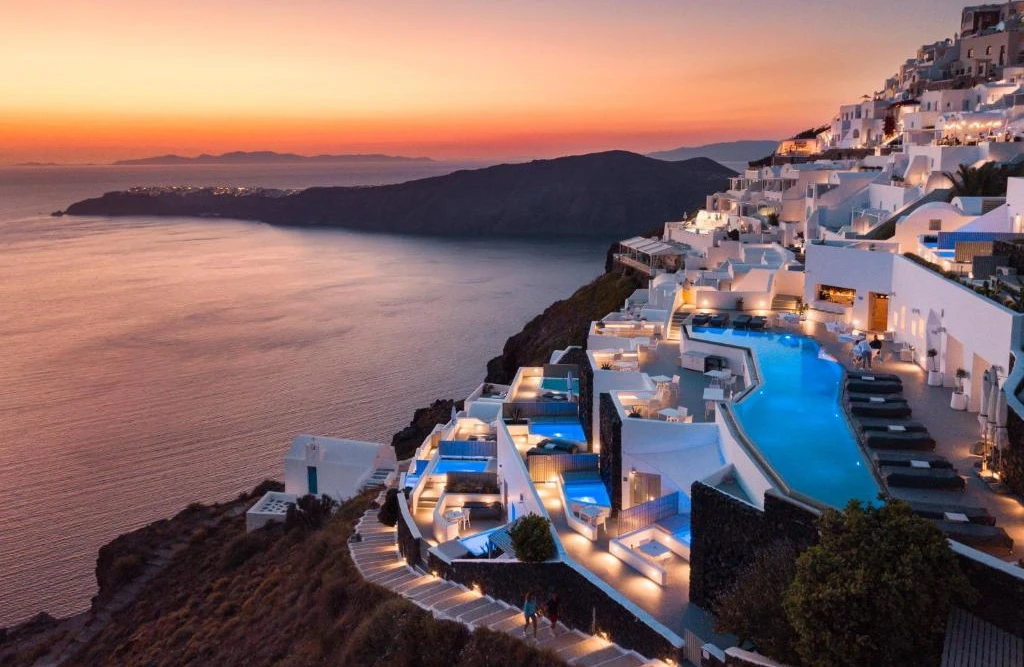 A photo of the Grace Hotel, a luxury hotel with a swimming pool, perched on a cliff overlooking the ocean at sunset. Santorini, Greece is known for its dramatic cliffs, whitewashed houses, and beautiful sunsets.