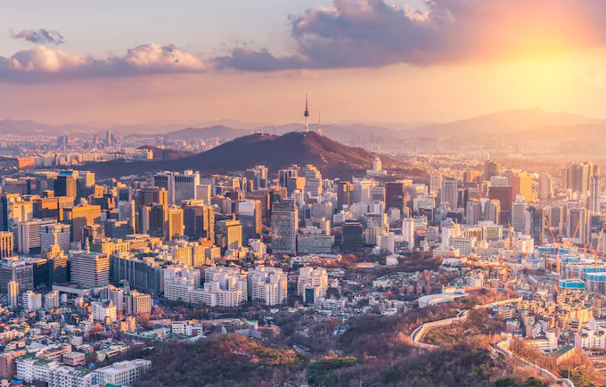 An aerial view of a cityscape at sunset. The city has tall buildings in a variety of shapes and sizes. There is a river or waterway winding through the city. In the background, there are mountains. [서울 Seoul South Korea]