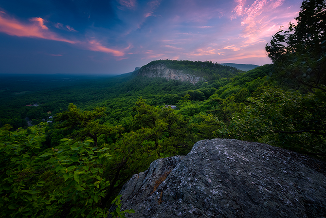 A scenic view of a Shawangunk mountain from the top of a mountain ridge. The valley floor is lush and green, filled with trees. The sky is a clear blue. In the distance, there are other, rolling hills and mountains.