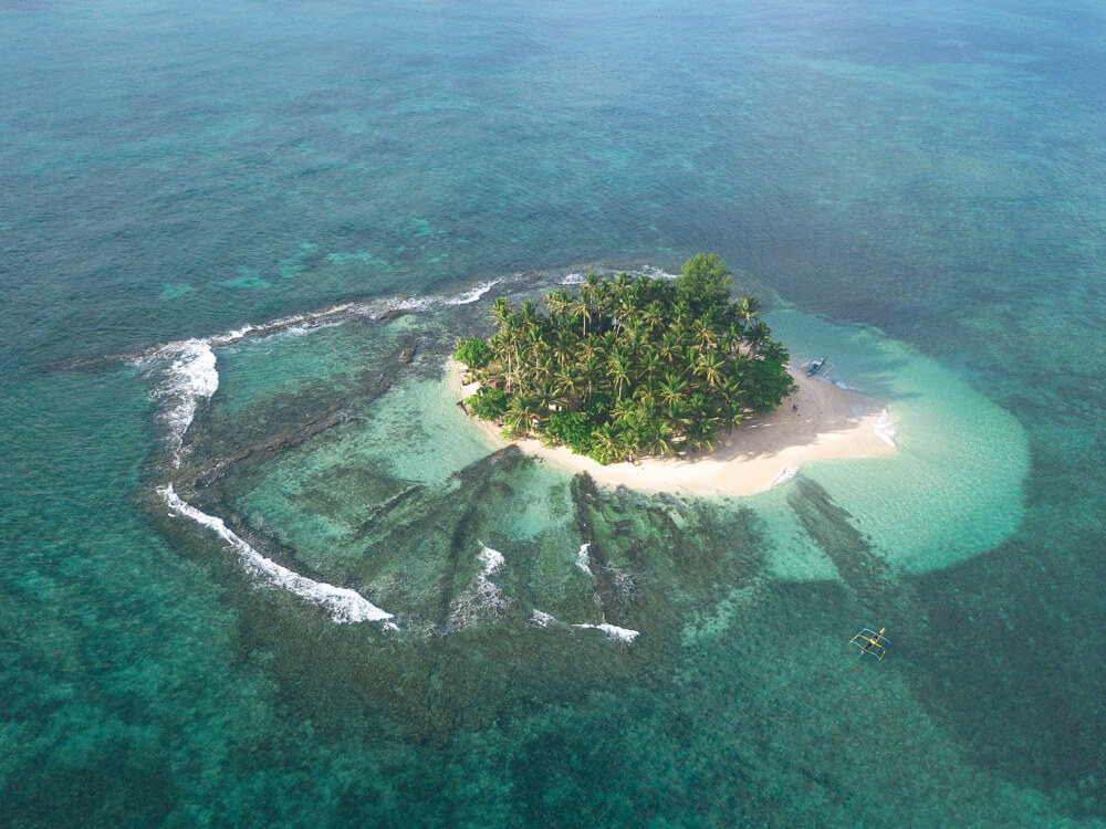 An aerial view of a small, tropical island surrounded by a blue ocean. The island has a white sand beach, palm trees, and lush vegetation.
