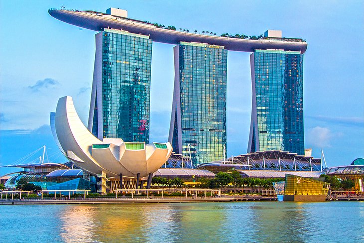 A photo of the Marina Bay Sands hotel complex in Singapore. The complex includes three skyscrapers connected by a горизонтальная платформенная конструкция (horizontal platform structure) with swimming pools and gardens. In the foreground is the ArtScience Museum, a lotus-shaped building.