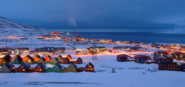 This image is of Svalbard Norway here is a row of houses in a snowy field at night. The houses are illuminated with warm yellow lights. The sky is dark and starry.