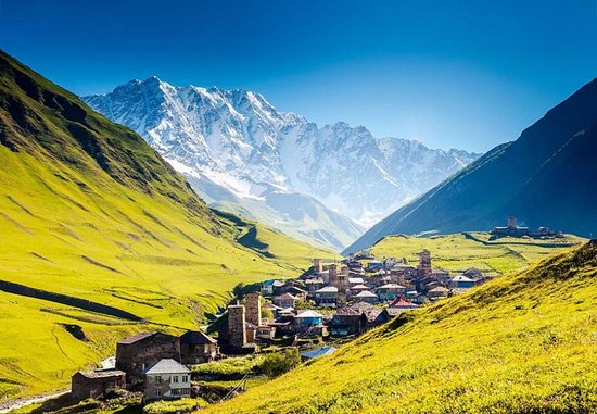 A photo of a village nestled at svaneti georgia in a lush green valley, surrounded by snow-capped mountains. The village has stone houses with slate roofs. A winding river flows through the valley.