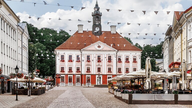 A photo of Tartu Town Hall, a red brick building with a tall tower and spire, located in Tartu, Estonia. The Town Hall is surrounded by a town square with trees and other buildings.