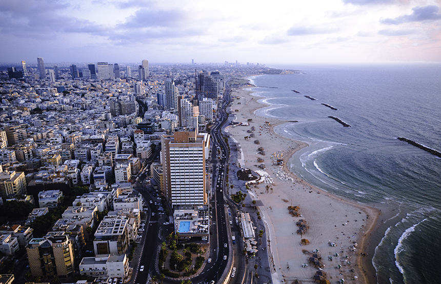 An aerial view of a city with a beach nearby. The city has skyscrapers and other buildings in various shapes and sizes. A winding river or waterway flows through the city and out to the ocean. The beach has white sand and calm, blue water. In the distance, there are mountains. [*[Tel Aviv, Israel]