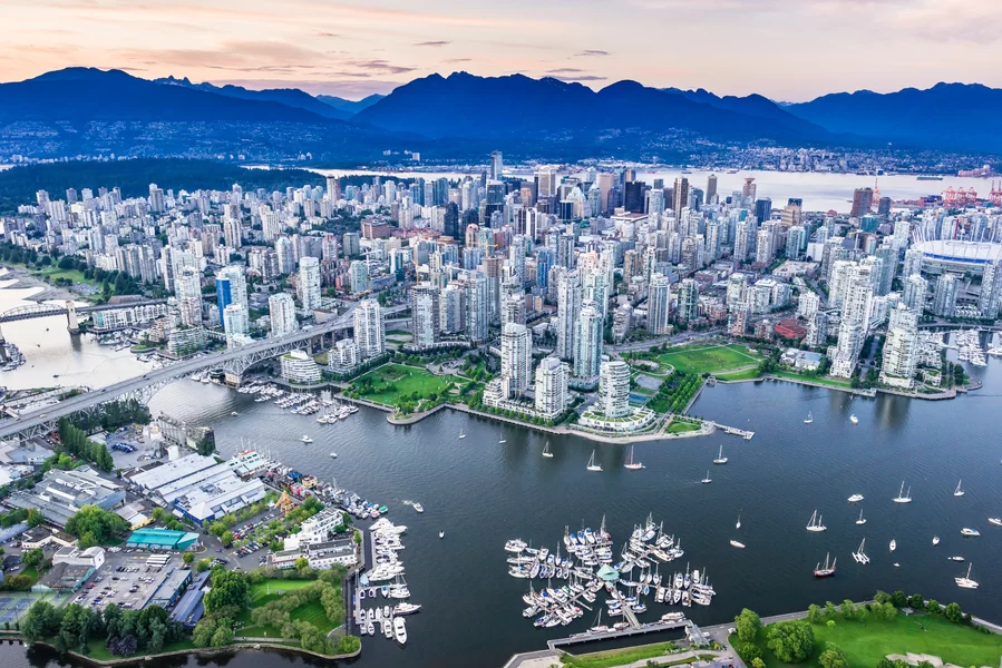 An aerial view of Vancouver, Canada, a city with a mix of modern skyscrapers and green spaces. Stanley Park, a large forested area, can be seen in the lower left corner. The Lions Gate Bridge can be seen in the distance, spanning the water between Vancouver and North Vancouver.