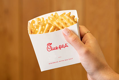 A hand holding a Chick-fil-A waffle fry container. The container is white with the Chick-fil-A logo and the phrase "sprinkled with sea salt." The fries are golden brown and appear crispy. Are Chick-fil-A Fries Gluten-Free