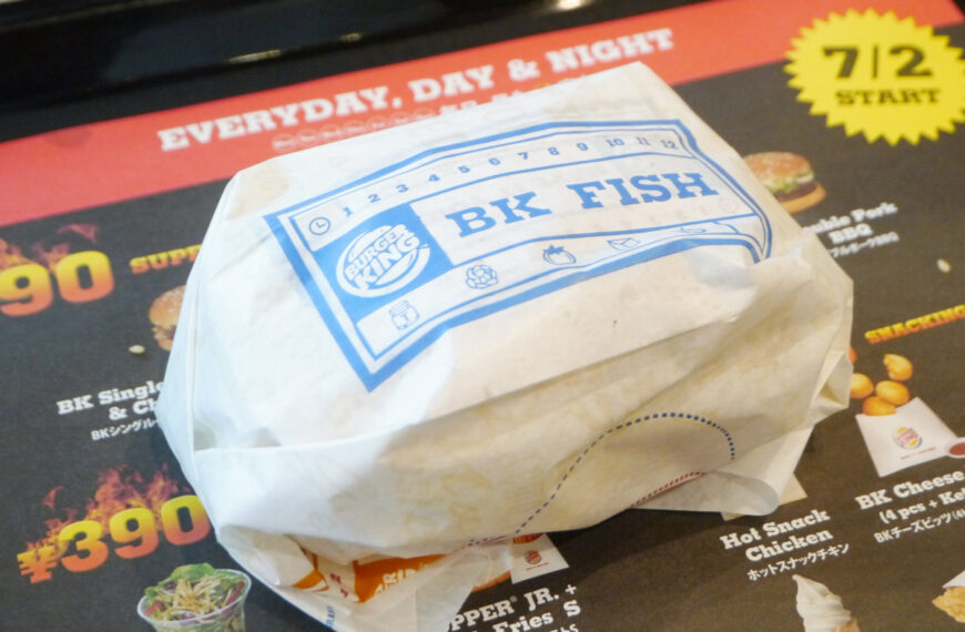 The image shows the burgerking fish sandwich in the warp on the table and behind it there is burger king menu also.