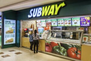 The picture shows the subway restaurant and some of ladys looking for the subway menu with prices.