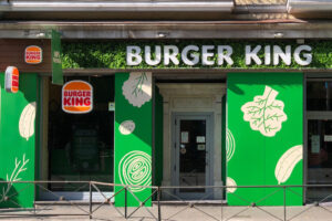 A green and white Burger King restaurant with a large, illuminated sign in the center. The restaurant features large, white leaf-shaped graphics and a green wall behind the sign. Discover the Burger King Breakfast Menu with Prices inside.