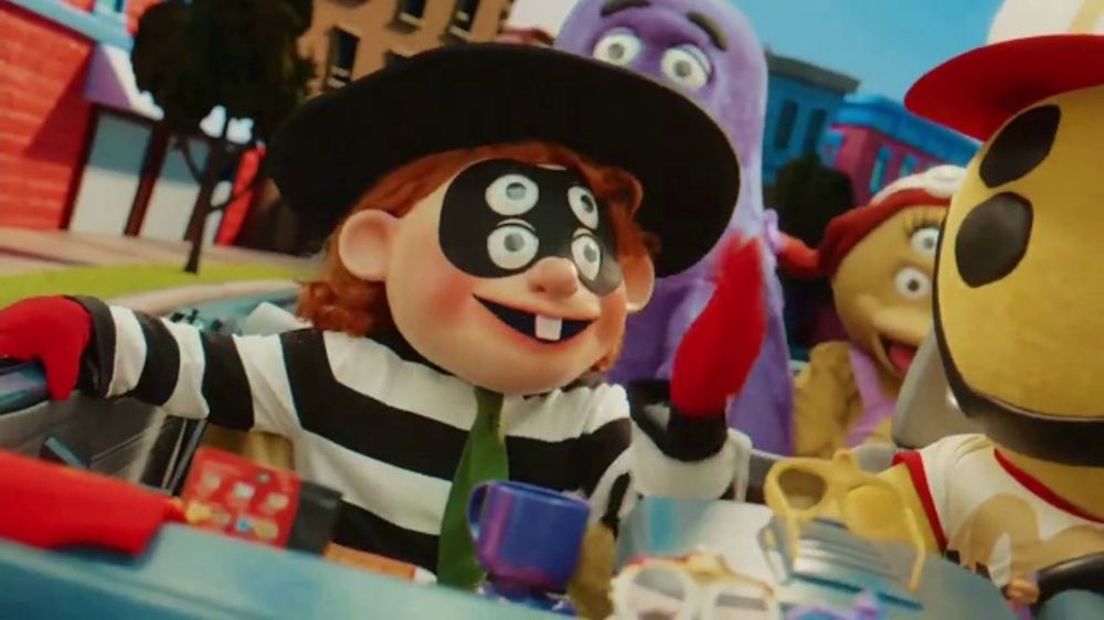 A cartoon character with a black and white striped outfit, a black hat, and three eyes is driving a car. The character has a wide smile and is waving to someone off-screen. There are other cartoon characters in the car with him, including a purple one and a yellow one.