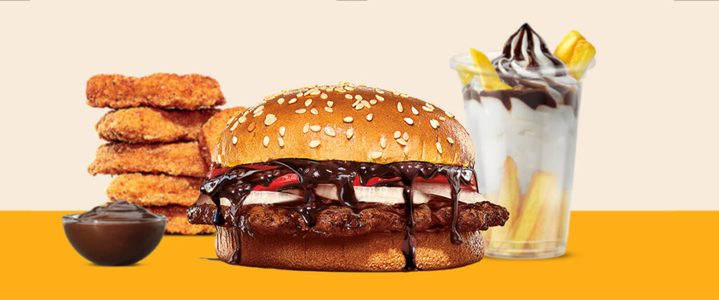  A Burger King meal featuring a chocolate Whopper, crispy chicken nuggets, fries, and a chocolate soft serve sundae. The Whopper has a sesame seed bun, a beef patty, and is drizzled with chocolate sauce.