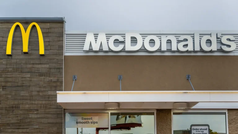 A photo of a McDonald's restaurant. The iconic golden arches logo is prominently displayed above the entrance. The restaurant's facade is made of brown brick, and the windows are large and inviting. A sign above the door reads "Sweet Smooth Sips," likely referring to the restaurant's beverage offerings. Another sign encourages customers to order ahead through the app. is McDonald’s Open On Labor Day