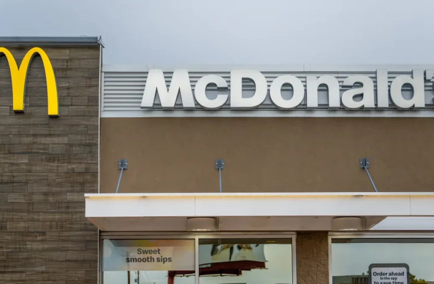 A photo of a McDonald's restaurant. The iconic golden arches logo is prominently displayed above the entrance. The restaurant's facade is made of brown brick, and the windows are large and inviting. A sign above the door reads "Sweet Smooth Sips," likely referring to the restaurant's beverage offerings. Another sign encourages customers to order ahead through the app. is McDonald’s Open On Labor Day