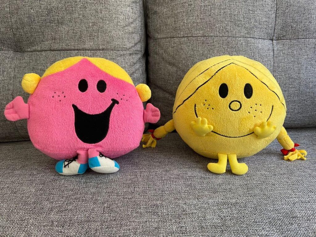 Two plush toys of Mr. Men characters. The pink one is Little Miss Sunshine, and the yellow one is Mr. Happy. They are sitting side-by-side on a gray couch.