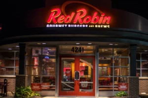 A brightly lit Red Robin restaurant with a large, red neon sign. The restaurant's facade is made of brick and glass, with double doors leading to the entrance. A small sign above the doors indicates that pickup orders are available. Red Robin Menu with Prices pdf usa