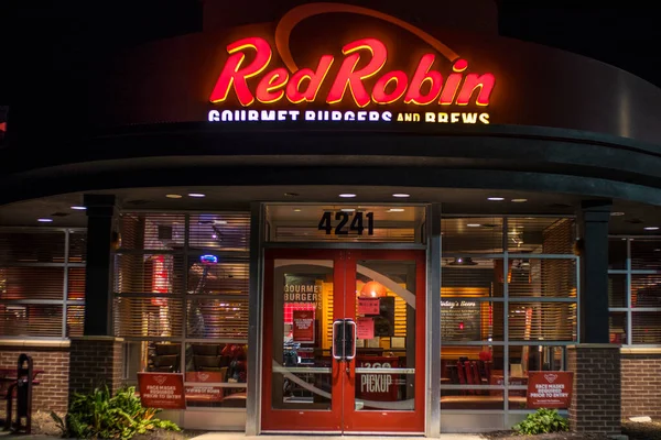 A brightly lit Red Robin restaurant with a large, red neon sign. The restaurant's facade is made of brick and glass, with double doors leading to the entrance. A small sign above the doors indicates that pickup orders are available. Red Robin Menu with Prices pdf usa