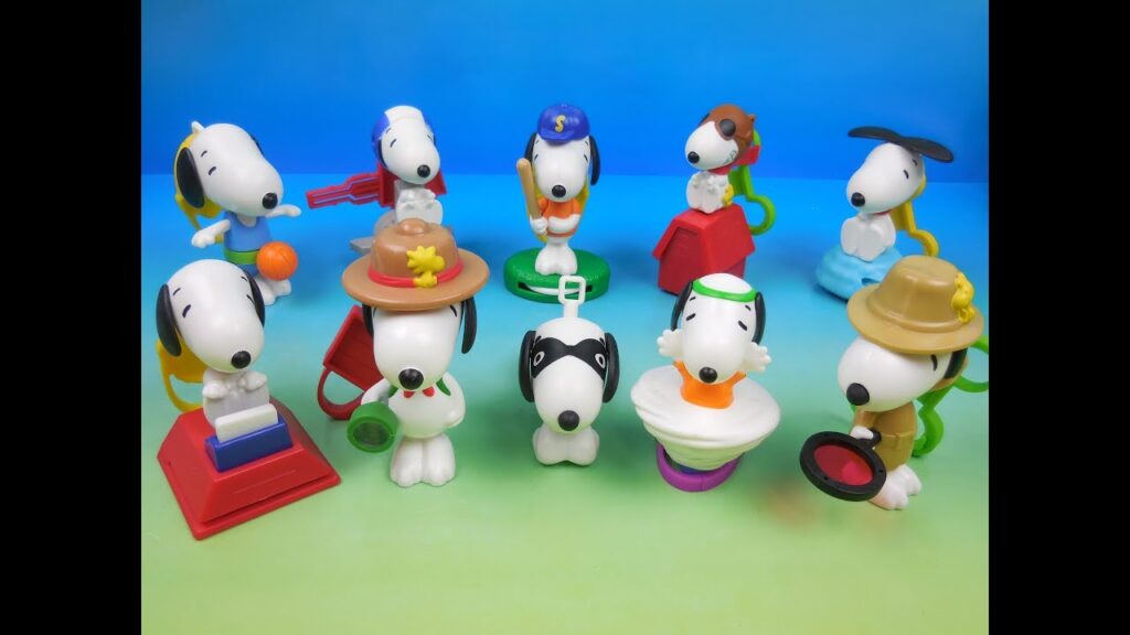 A collection of colorful plastic toys featuring Snoopy and his friends from the Peanuts comic strip. The toys depict Snoopy in various costumes and activities, including playing baseball, wearing a police hat, and being a firefighter. The toys are arranged on a green and blue background.