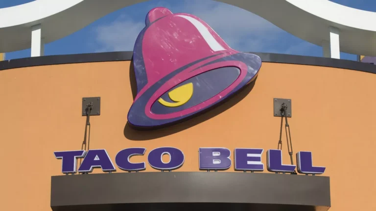 A Taco Bell restaurant sign featuring the signature purple and pink bell logo, with the words "Taco Bell" displayed below. Take advantage of Taco Bell Deals Box for exclusive offers!