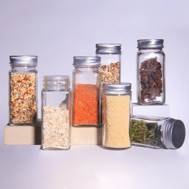  A set of glass spice jars with silver lids. The jars are filled with various spices and herbs, such as red pepper flakes, star anise, and dried herbs.