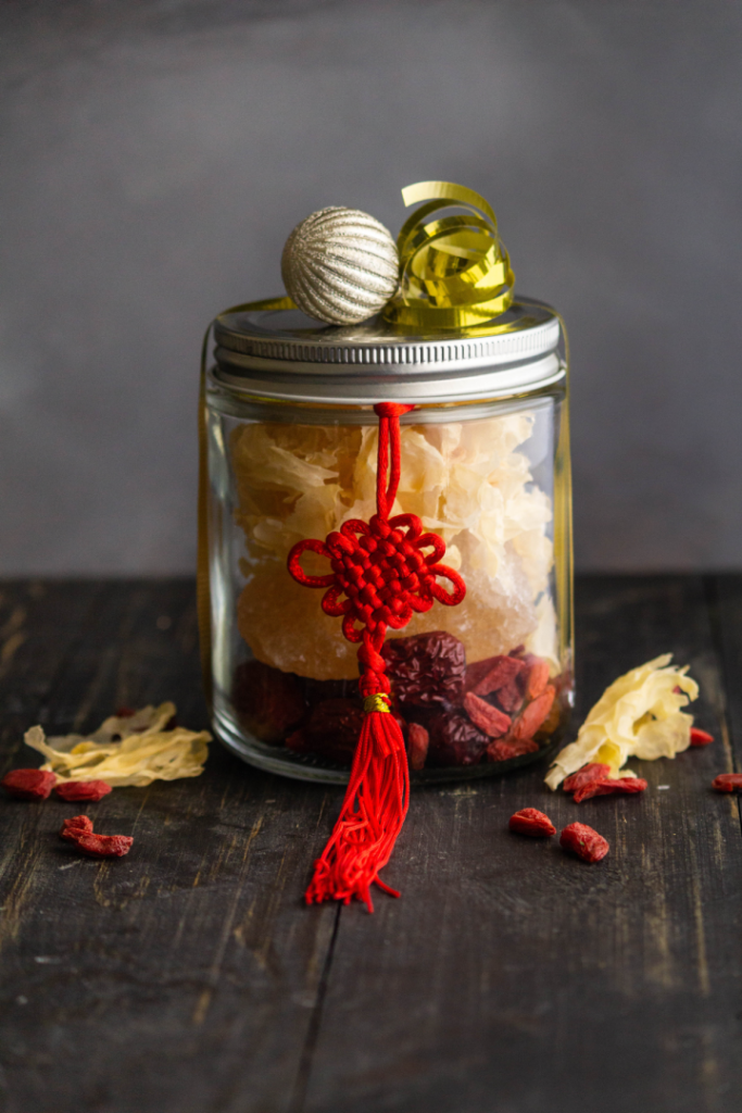 A glass jar filled with dried white fungus and goji berries. A red Chinese knot is tied around the jar, and a silver ornament and gold ribbon decorate the top.