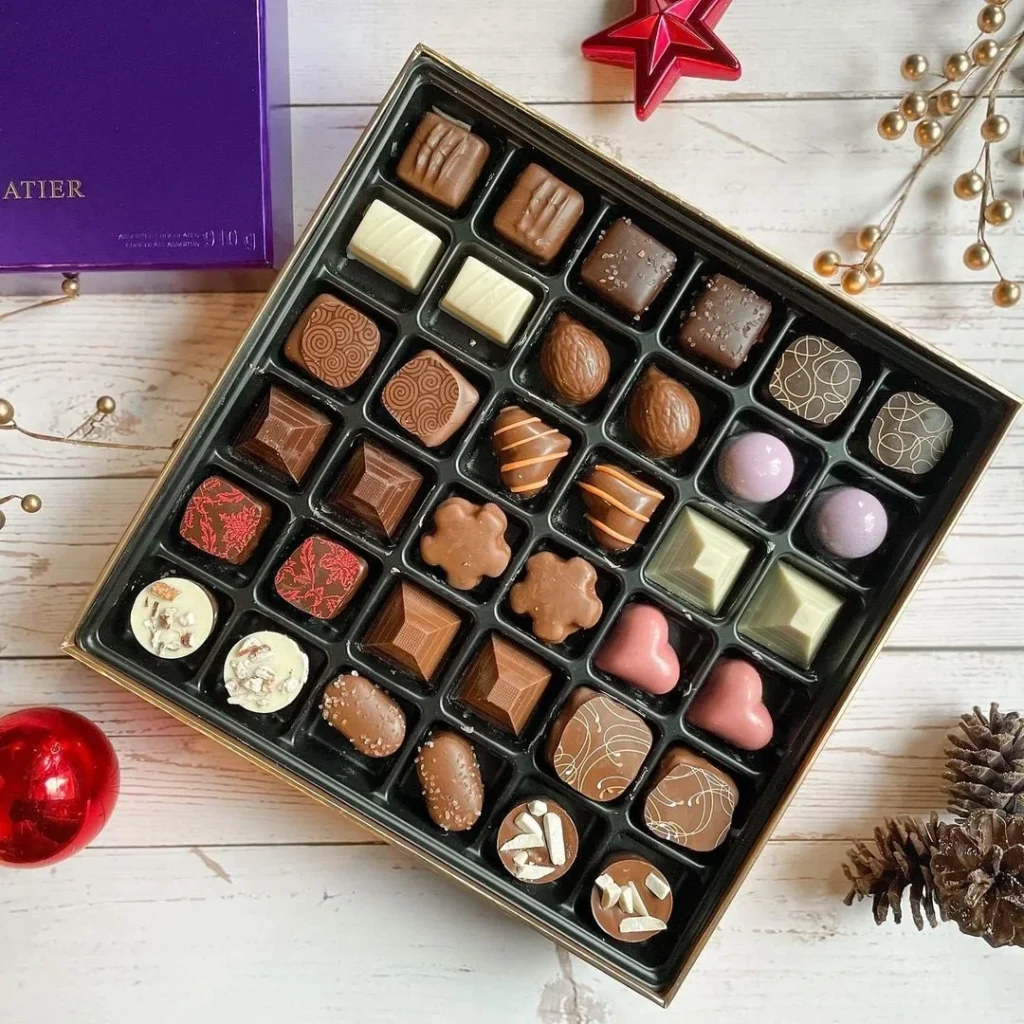 A luxurious box of assorted chocolates from Atelier de la Table. The chocolates are arranged in compartments of various shapes and sizes, and the box is surrounded by festive decorations like a red star and pine cones.