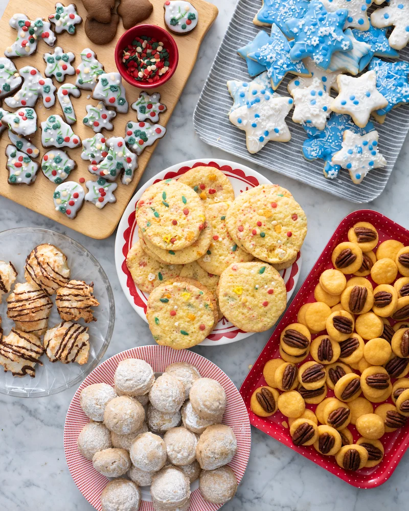  A variety of Christmas cookies arranged on multiple plates and trays. There are sugar cookies decorated with frosting and sprinkles, pecan cookies, and other types of holiday treats.it is a Delicious Food Gifts for Christmas