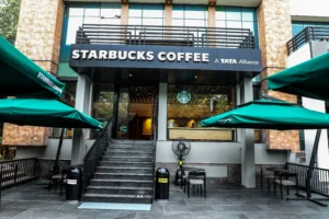 A Starbucks cafe exterior with green umbrellas and outdoor seating. The sign above the entrance reads "Starbucks Coffee, A Tata Alliance". This location offers a variety of drinks, including those listed on the Starbucks Menu With Calories.